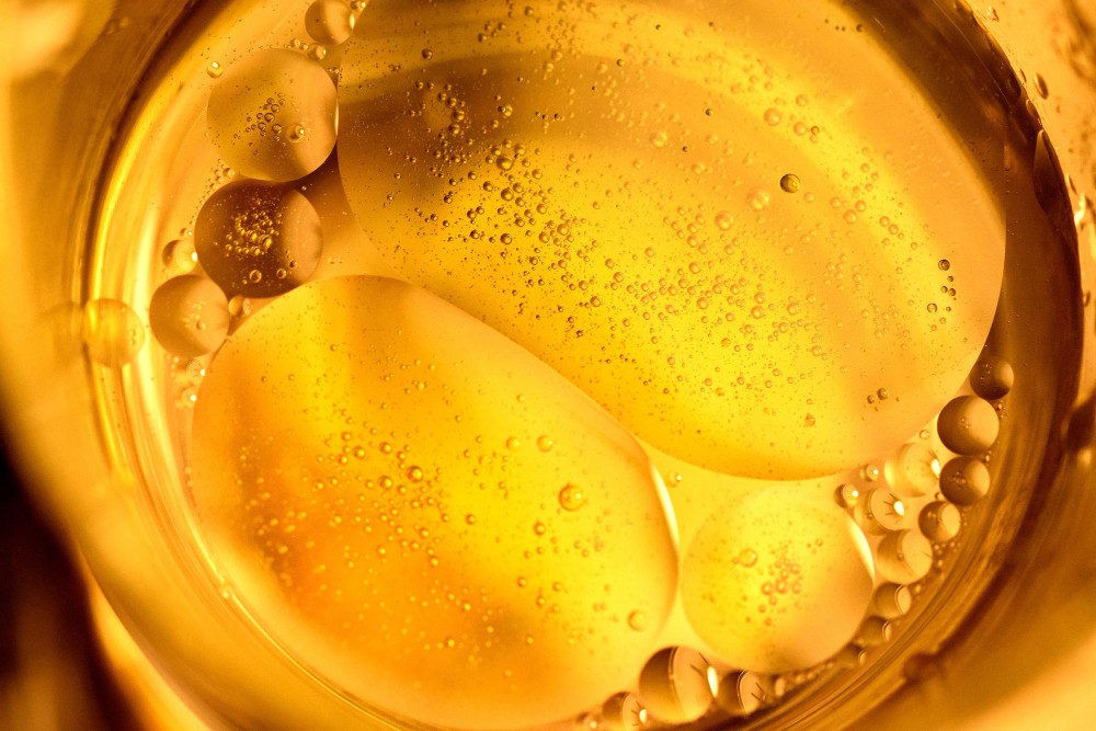 A close-up, top-down view of a golden-yellow, viscous liquid, resembling honey or oil, filled with numerous bubbles and larger, translucent pockets. The surface glistens with reflected light.