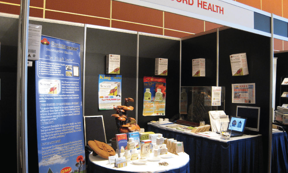 A well-lit trade show booth featuring black walls and a prominent blue and white banner with product information and a 'GMP' logo. A white circular table in the foreground displays an array of health product boxes and bottles, while behind it, dried Ganoderma mushrooms are exhibited. A rectangular table on the right holds more brochures and a laptop, with a partial 'HEALTH' sign visible overhead.