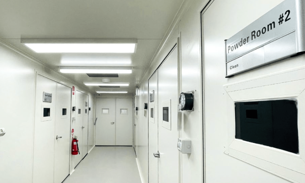 A brightly lit, sterile hallway with white walls and ceiling, typical of a cleanroom or industrial facility. Numerous white doors with small viewing panels line both sides of the corridor. A prominent sign on the right wall reads 'Powder Room #2' with 'Clean' underneath. A fire extinguisher is mounted on the left wall, and a circular pressure gauge is visible on one of the doors on the right.