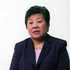 A middle-aged East Asian woman with short, dark hair is shown from the chest up, wearing a dark pinstriped suit jacket over a light-colored top and a necklace. Her mouth is slightly open as if speaking, indicating she is giving a testimonial. She is positioned against a plain white background.