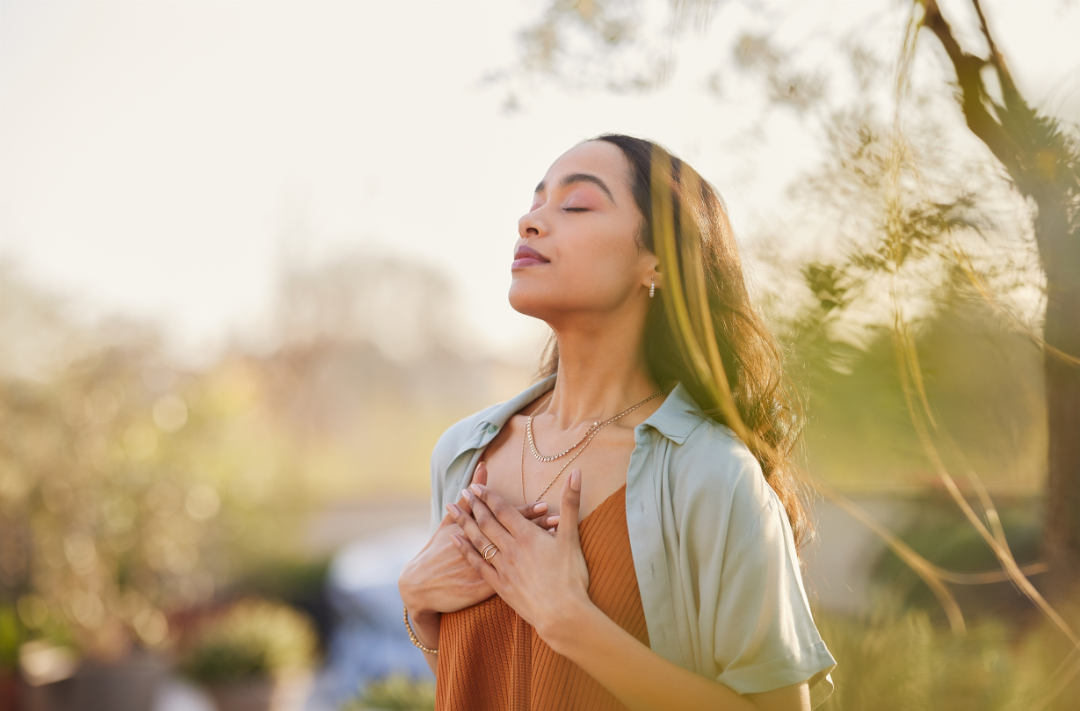 A woman with long, flowing dark hair stands outdoors with her eyes closed and head tilted slightly upward, hands gently clasped over her chest. She wears a rust-colored top and a light green unbuttoned collared shirt. The background is a softly blurred blend of green and yellow foliage, suggesting a peaceful, sunny natural environment.