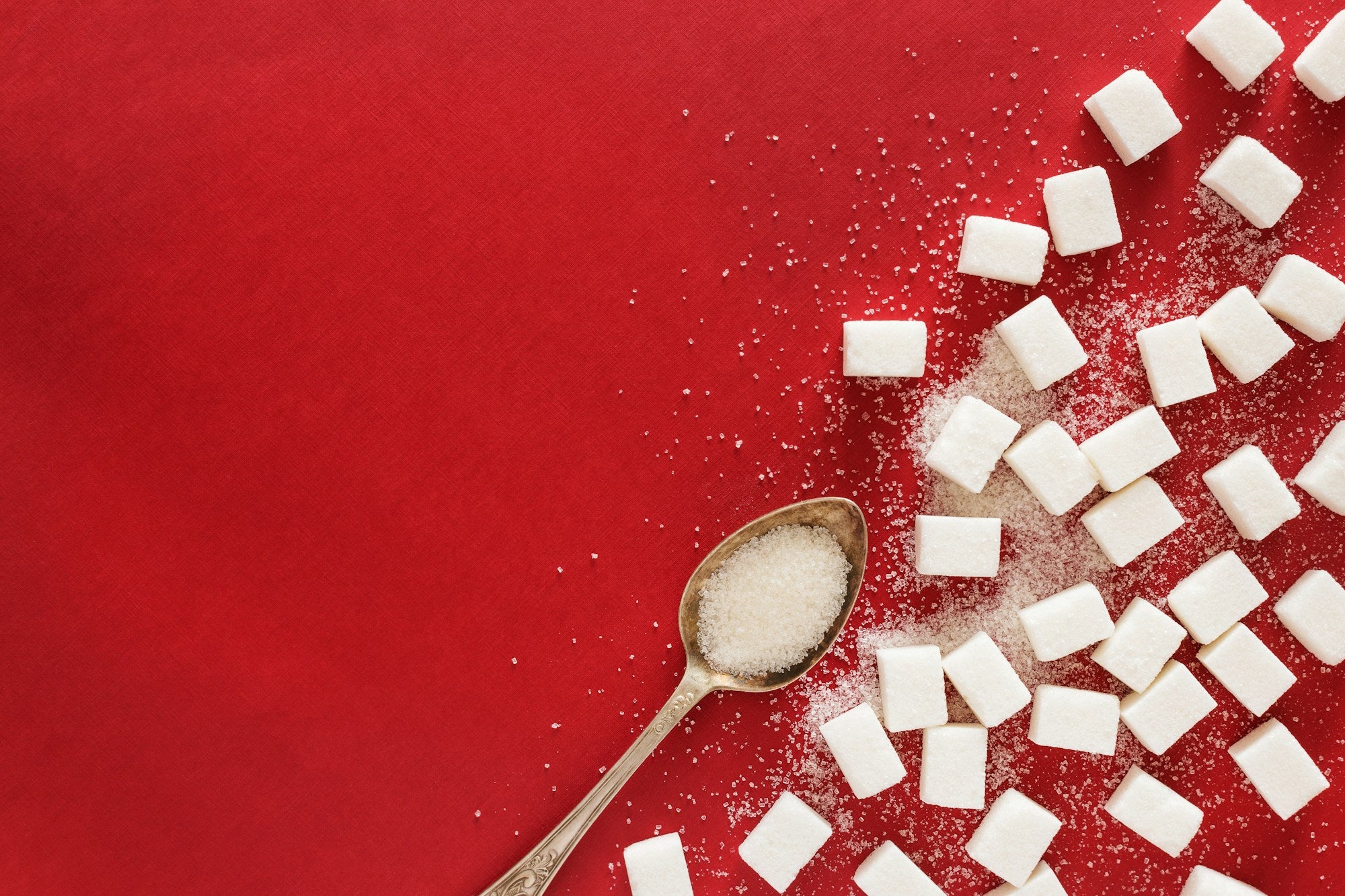 A top-down view of a vibrant red surface with white sugar cubes and loose granulated sugar scattered on the right side. A silver spoon with an ornate handle rests on the red background towards the bottom left, holding a small amount of granulated sugar in its bowl.