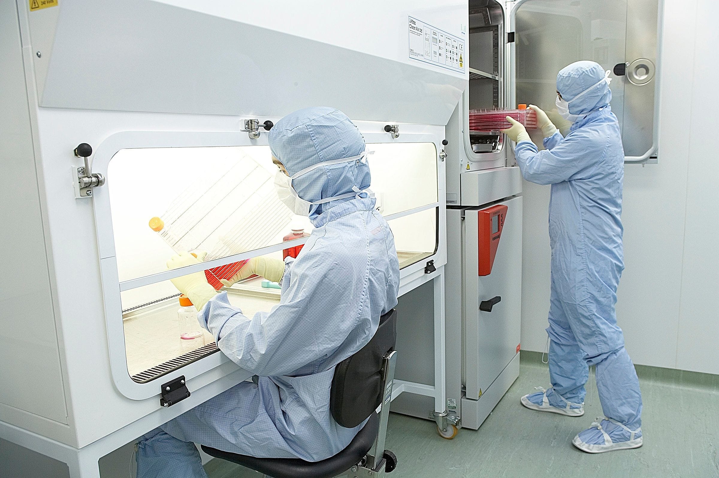 Two individuals in full light-blue cleanroom suits, masks, and gloves are working in a sterile laboratory. One person is seated at a brightly lit laminar flow hood, with their arms extended into the workspace. The second person stands upright, placing a multi-well plate into a large, open, white laboratory incubator or storage unit. The overall environment is very clean and organized.